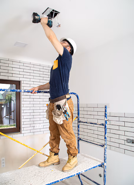 eletricista-construtor-em-obra-instalacao-de-lampadas-em-altura-profissional-de-macacao-com-uma-broca-no-fundo-do-local-de-reparo_169016-8248